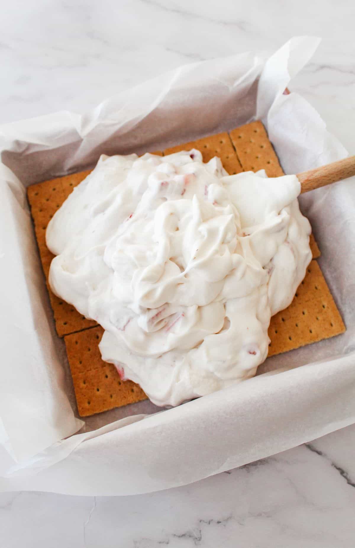 Ice cream mixture being spread out onto graham crackers in a parchment lined baking dish. 