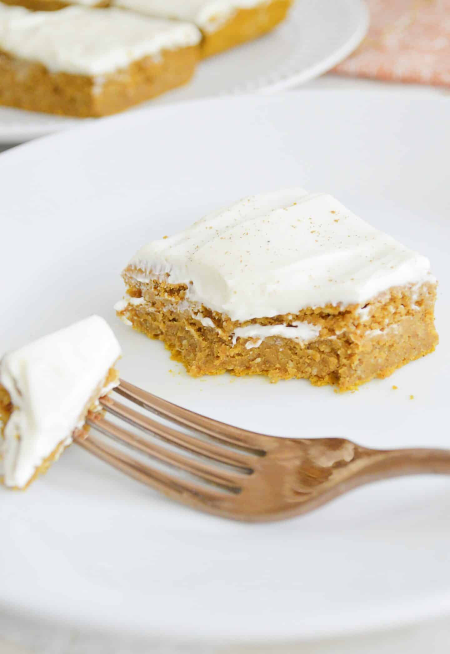 Pumpkin bar with cream cheese frosting on a white plate with a bite taken out of it. 