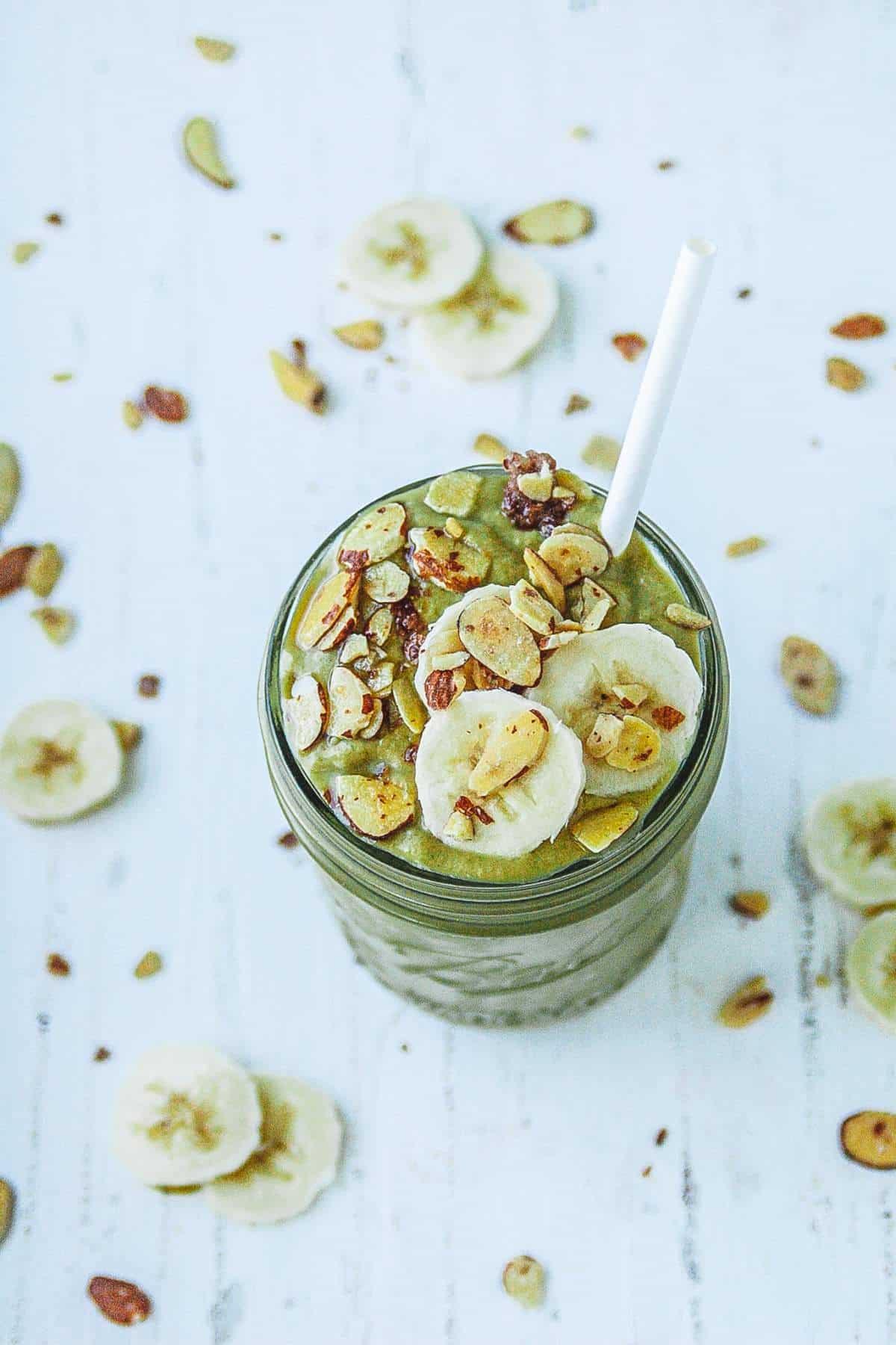 Smoothie with mango, spinach, and banana in a mason jar with banana and almonds spread around it. 