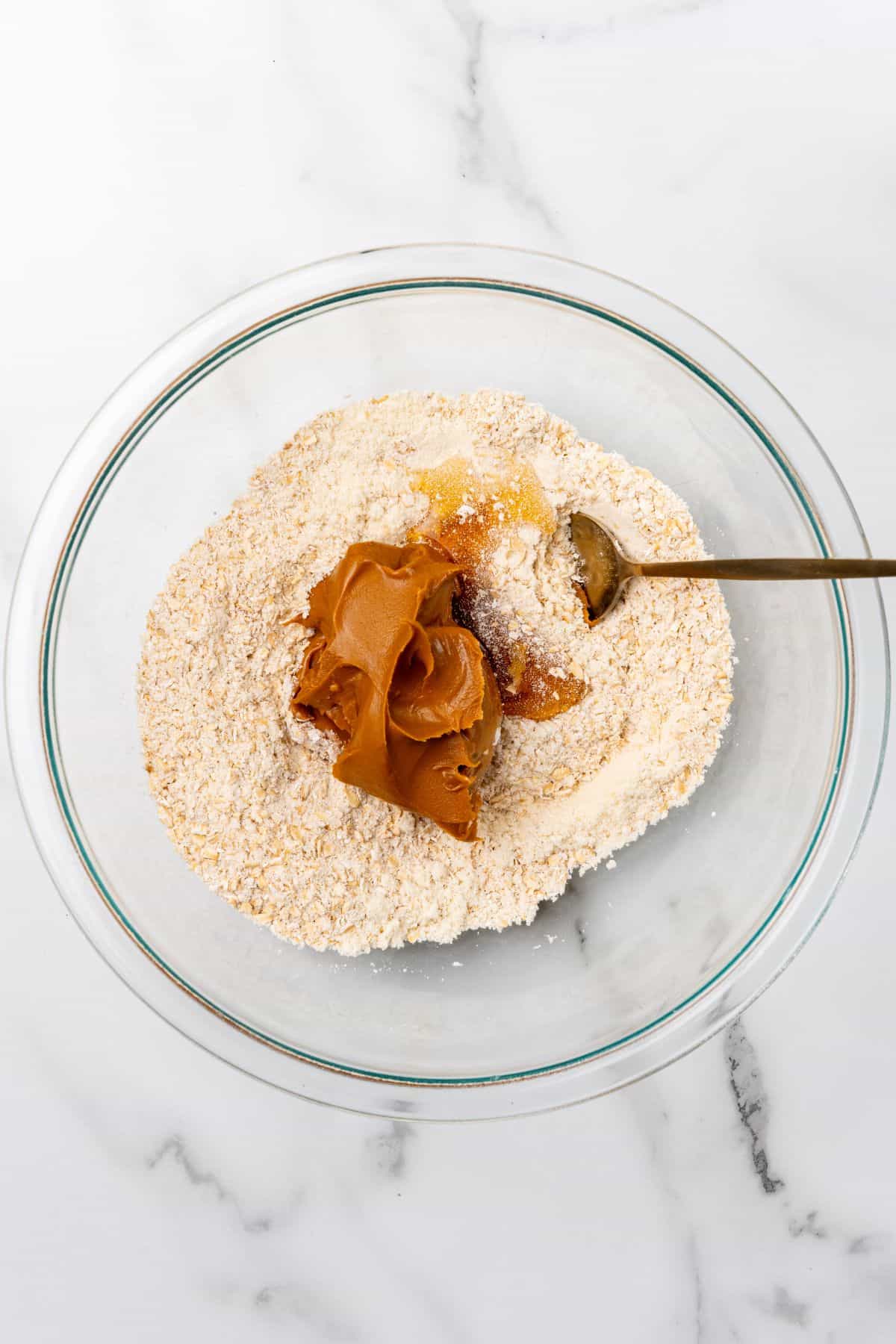 A glass bowl with oats, protein powder, Biscoff, and honey being mixed together. 
