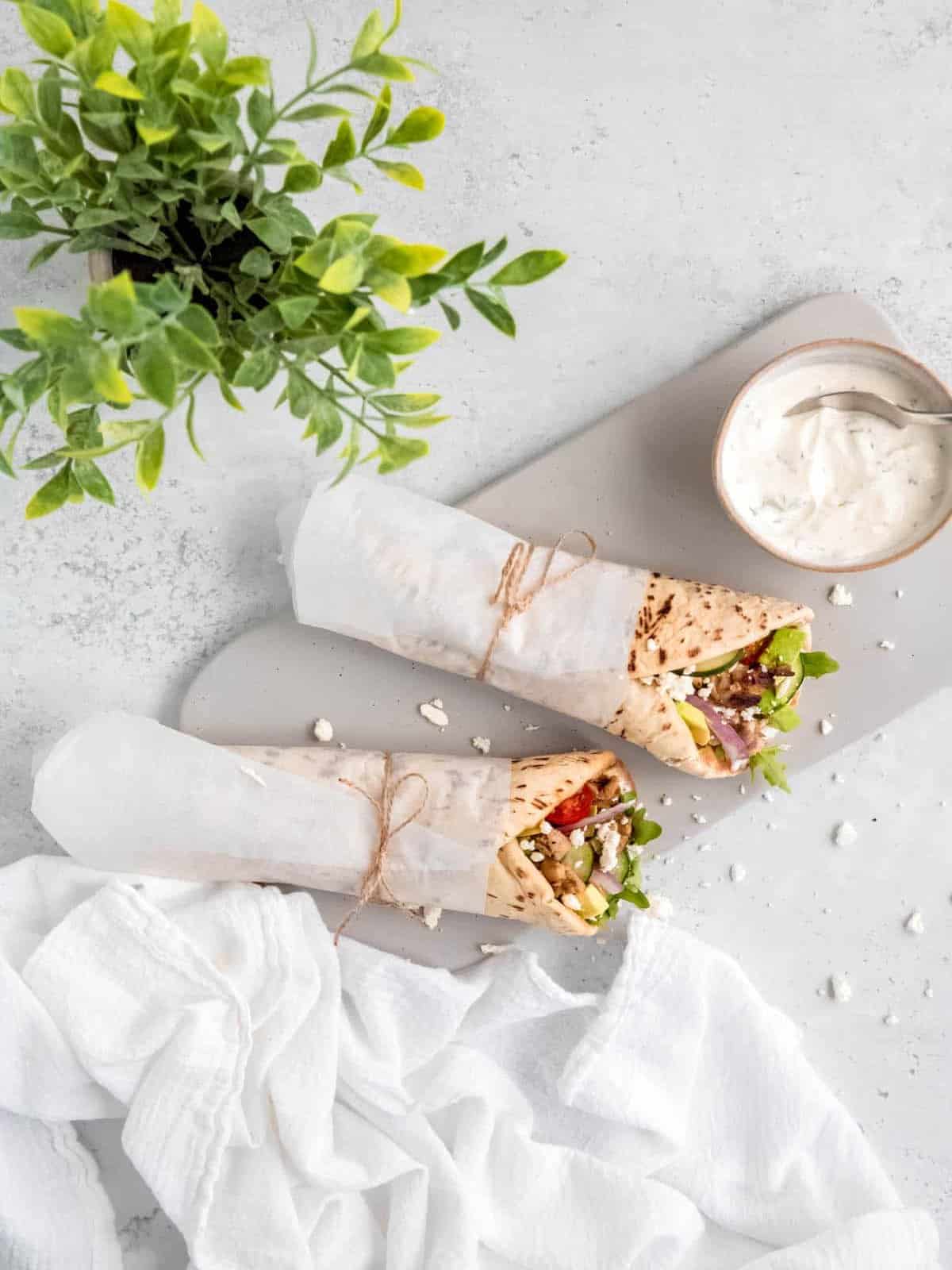 Grilled chicken pita wraps on a white plate with with Tzatziki sauce in a bowl and a plant in the background.