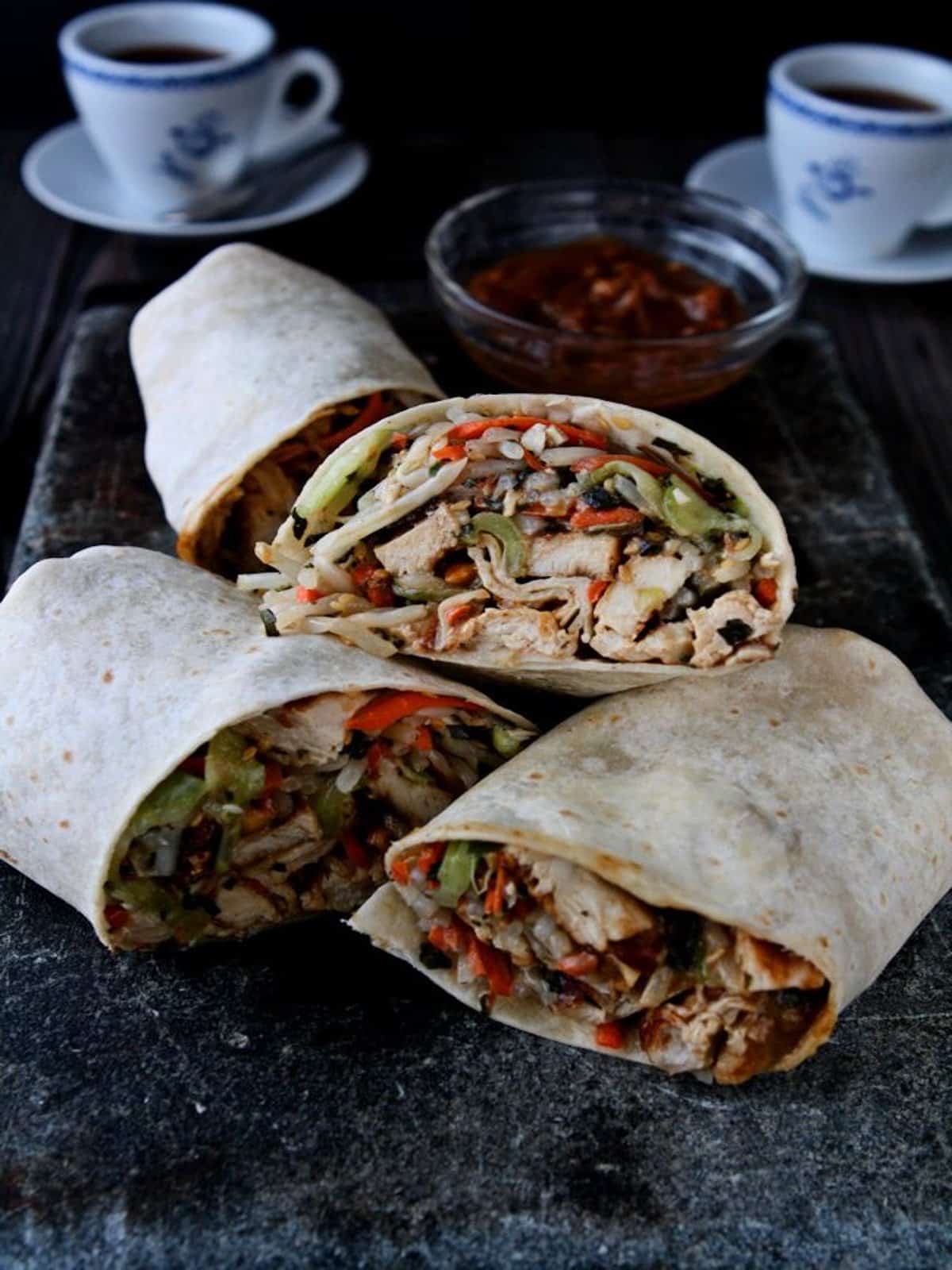 Three Thai chicken wraps on a platter with a bowl of peanut sauce. 