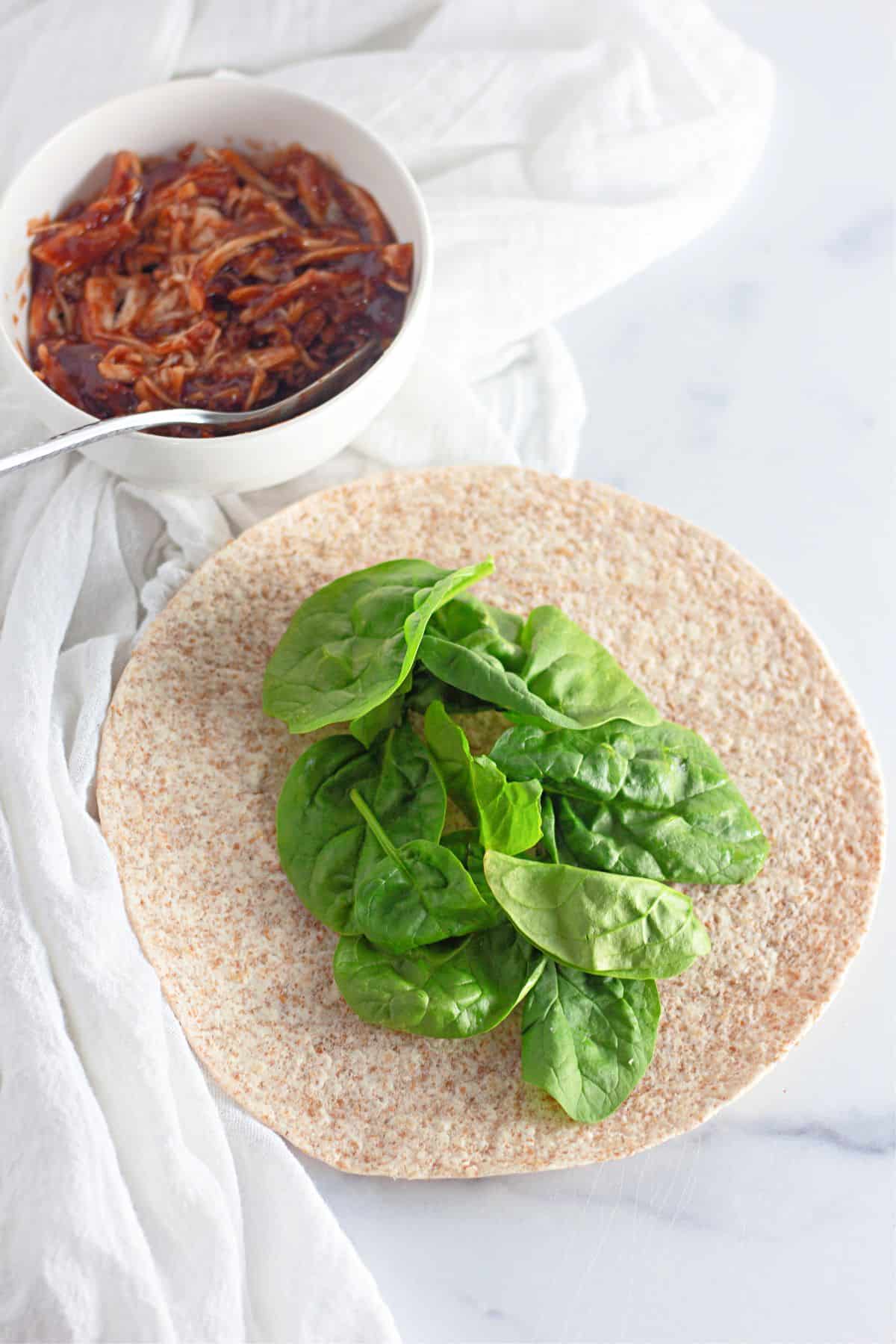 A wrap with spinach and a bowl of BBQ chicken.