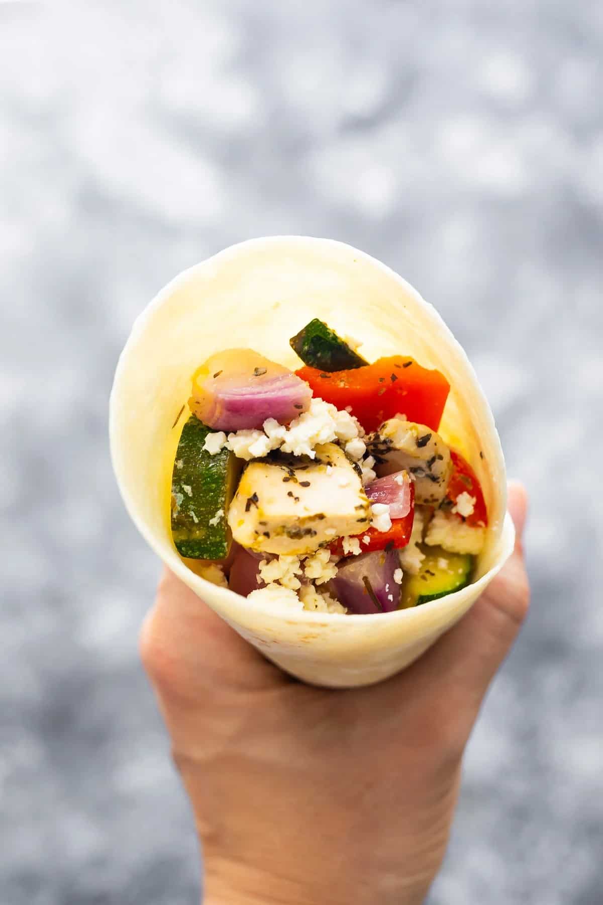 A person is holding a Greek chicken wrap with feta cheese roasted vegetables in it.