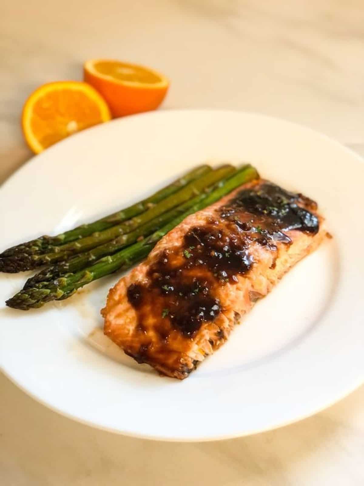 Glazed salmon and asparagus served on a white plate with an orange next to it. 