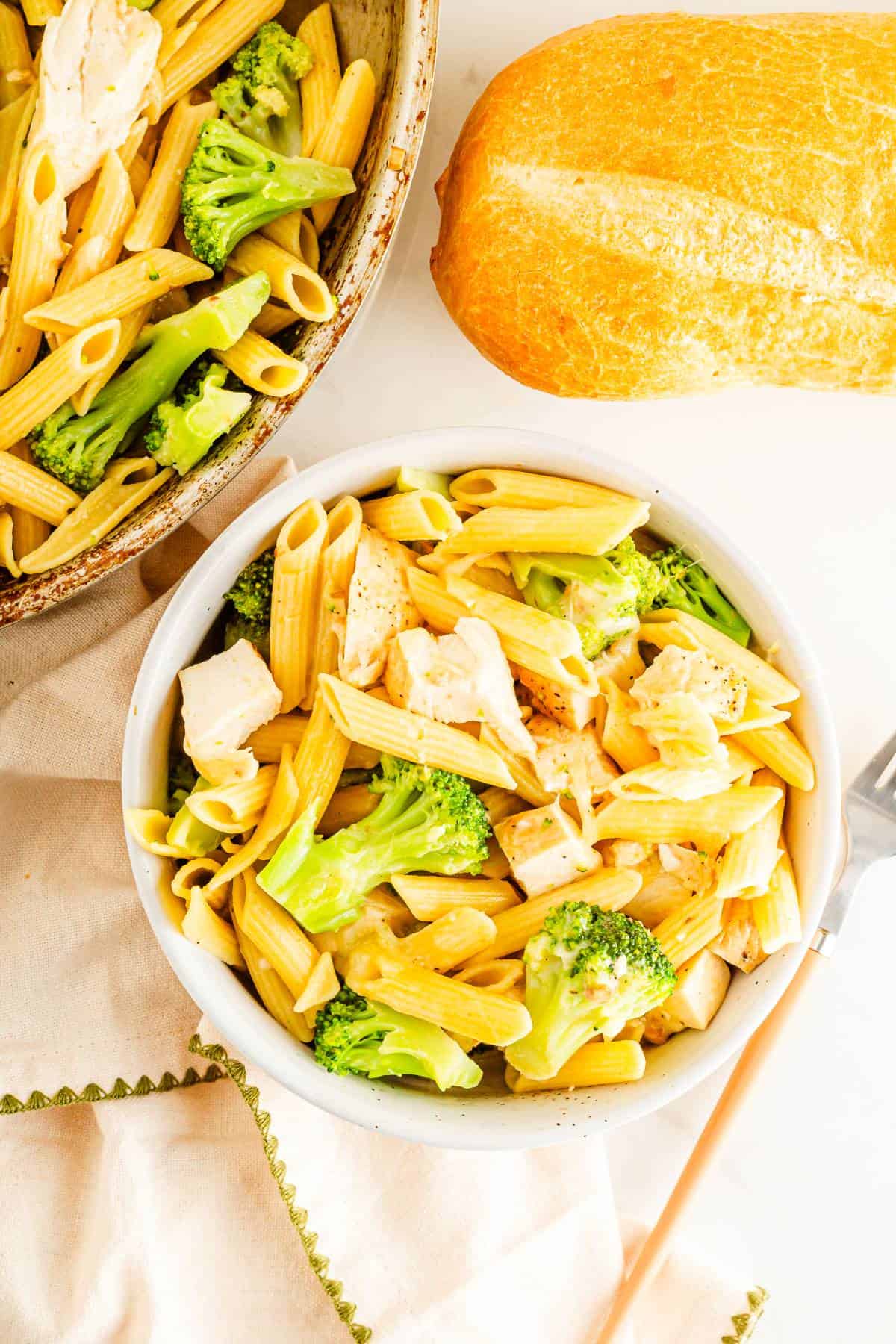 A bowl of penne pasta with chicken and broccoli being served with bread. 