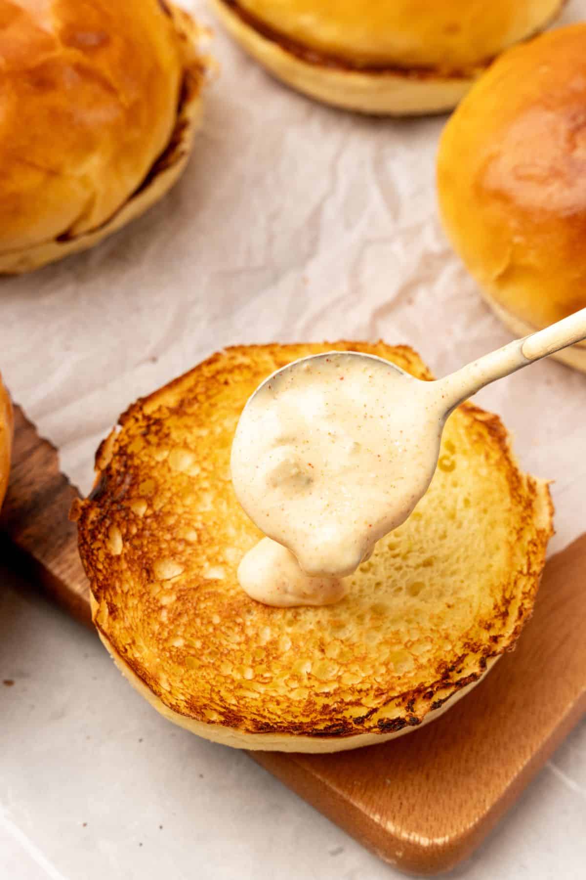 A spoon is drizzling smash sauce on a toasted bun. 