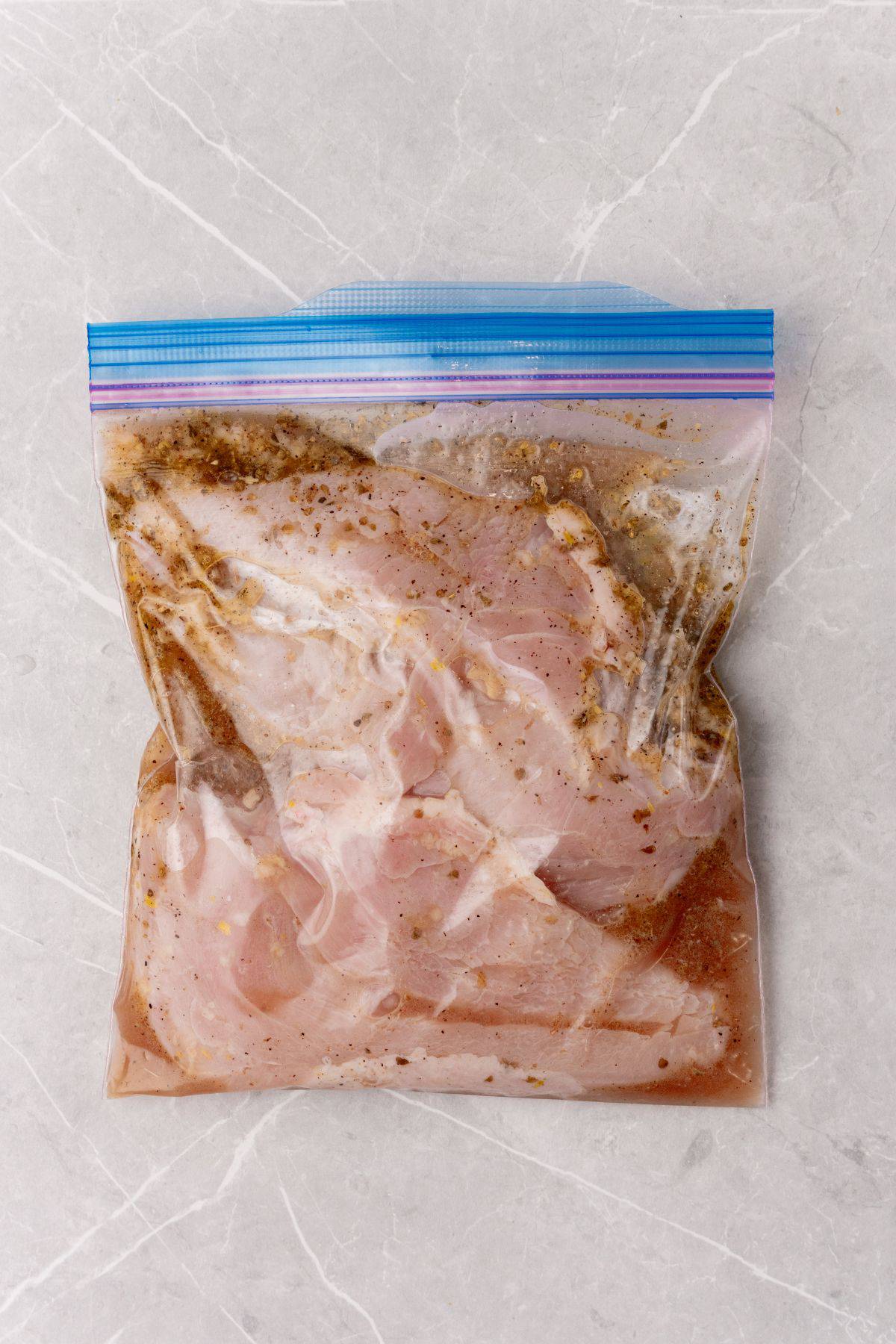 A plastic bag filled with in marinated chicken on a marble surface.