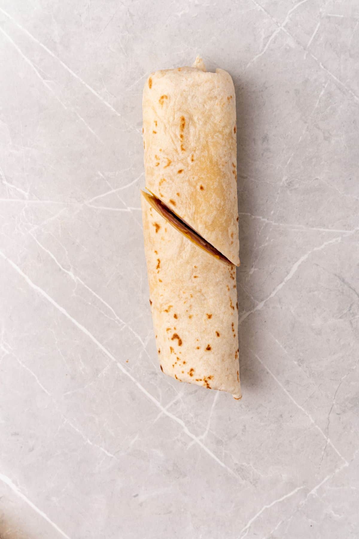 A cheese-filled burrito wraps generously with slices of ham on a sleek marble surface.