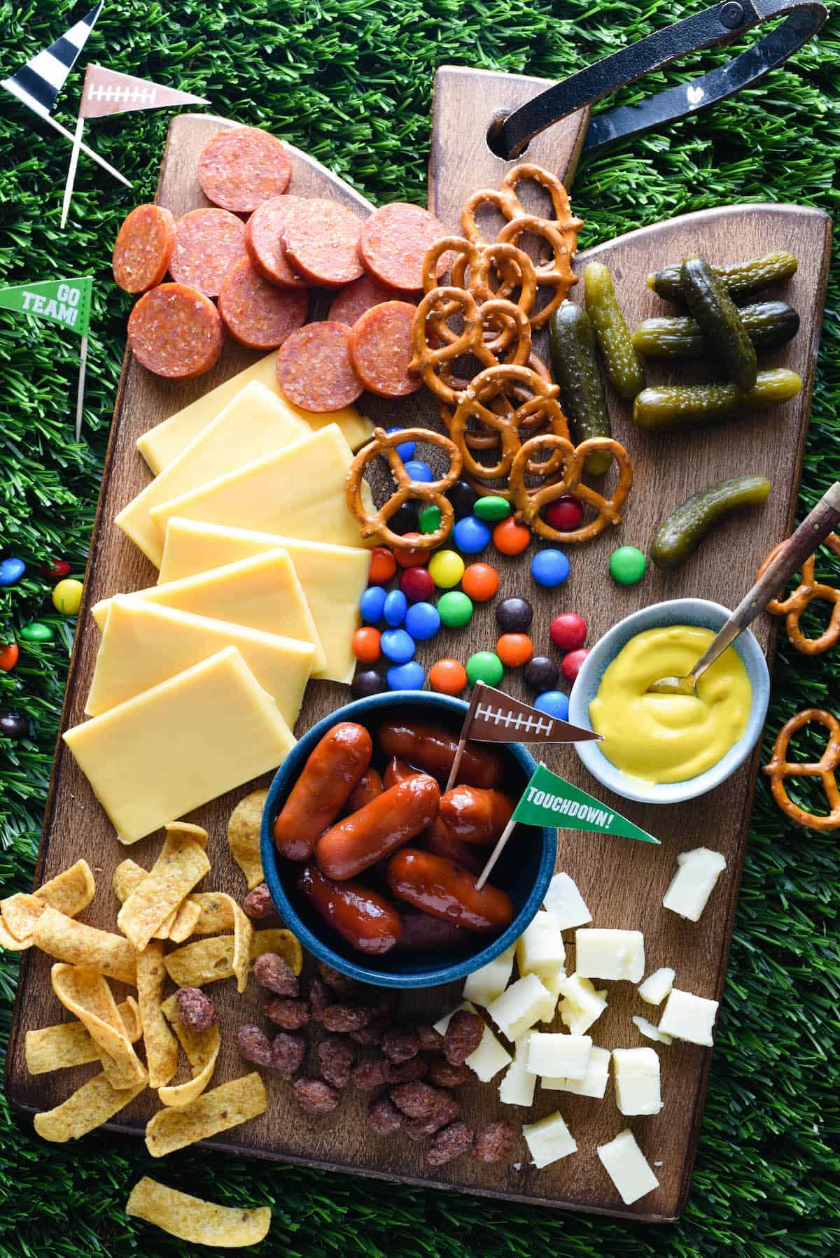 Assorted snacks on a wooden board including cheese, sausages, pretzels, pickles, and dips, arranged for a sports-themed party.