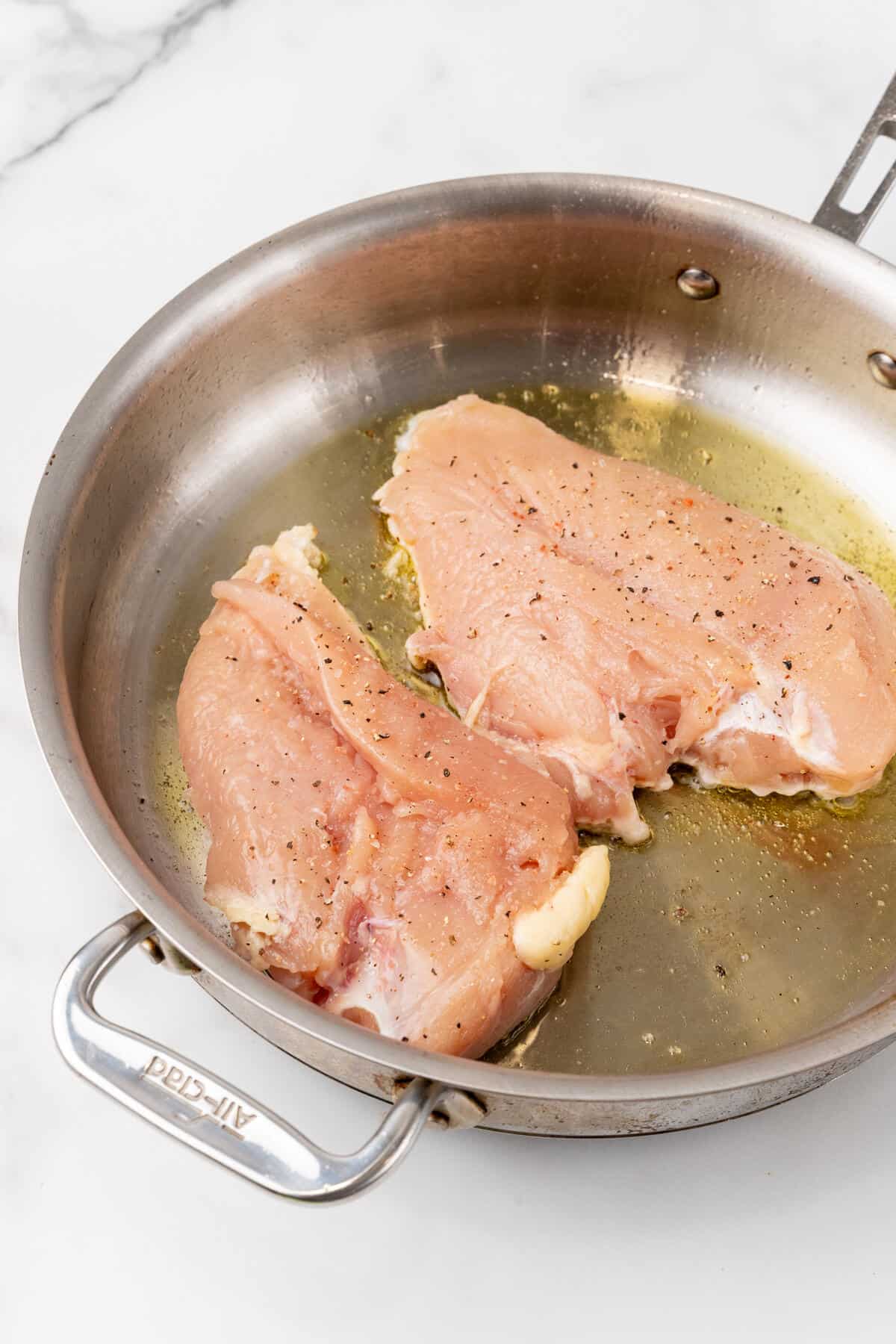 Two chicken breasts being sautéed in a stainless steel pan.