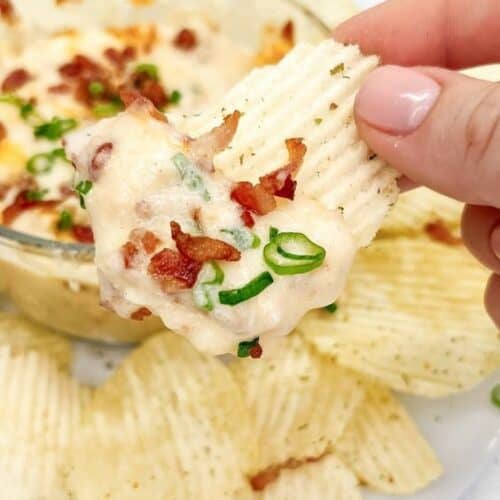 A hand holding an appetizer chip with creamy dip and toppings of bacon and green onions.