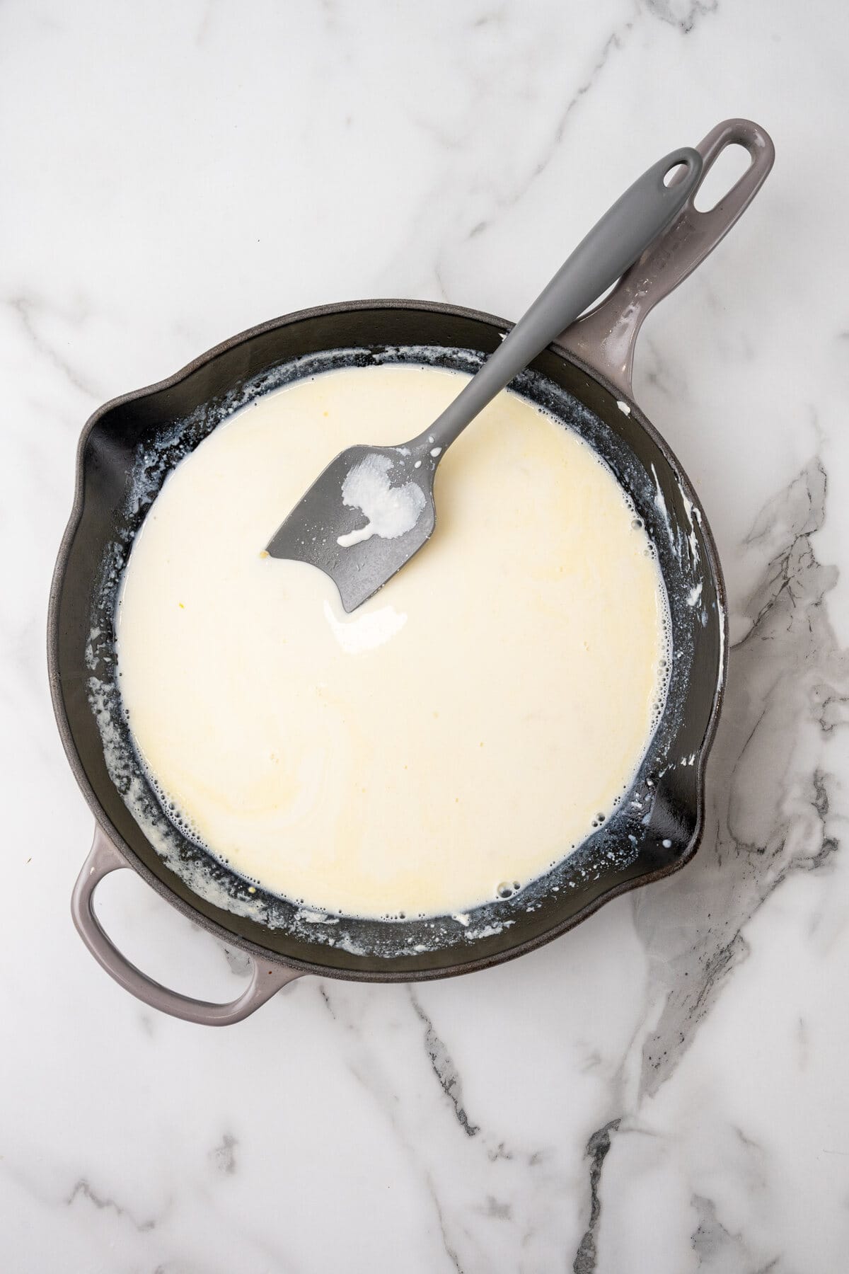 A cast iron skillet with melted butter, milk, and a spatula on a marble countertop.