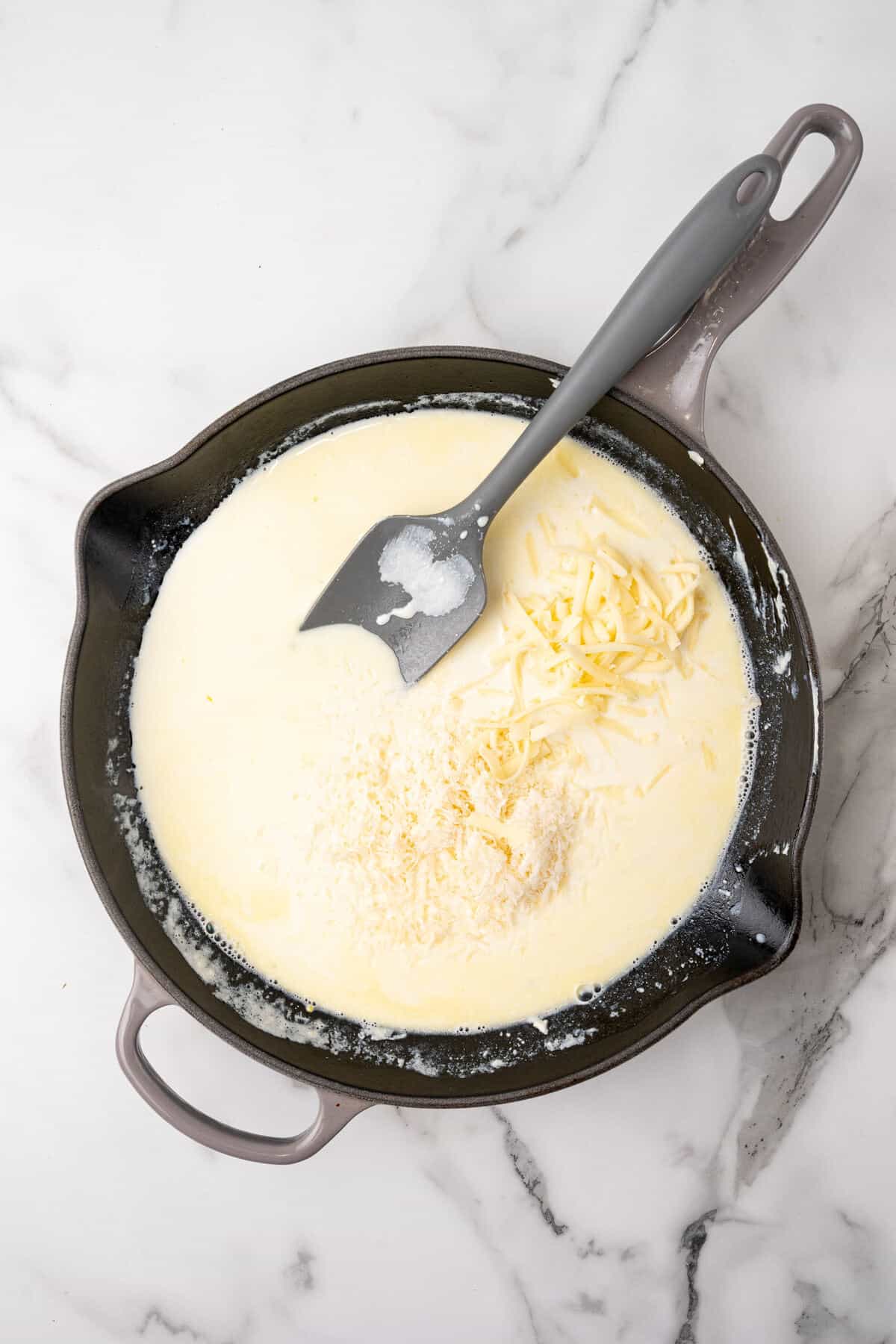 A skillet containing a creamy sauce and cheese, alongside a spatula on a marble countertop.