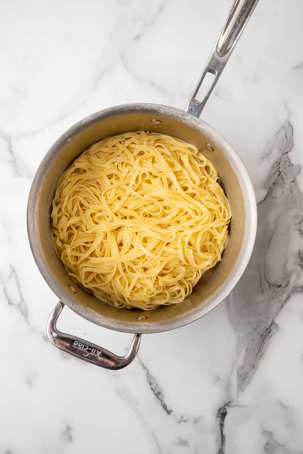 Cooked spaghetti in a stainless steel pot on a marble countertop for an Alfredo recipe.