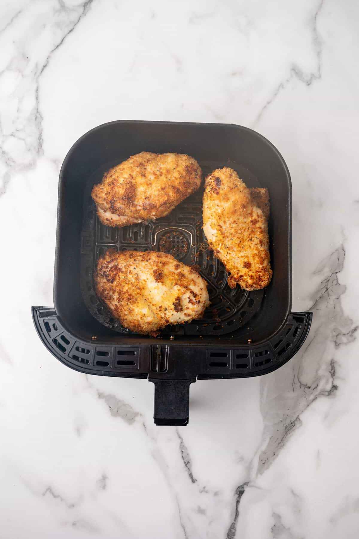 Three breaded Parmesan chicken breasts cooking in a air fryer basked on a marble surface.
