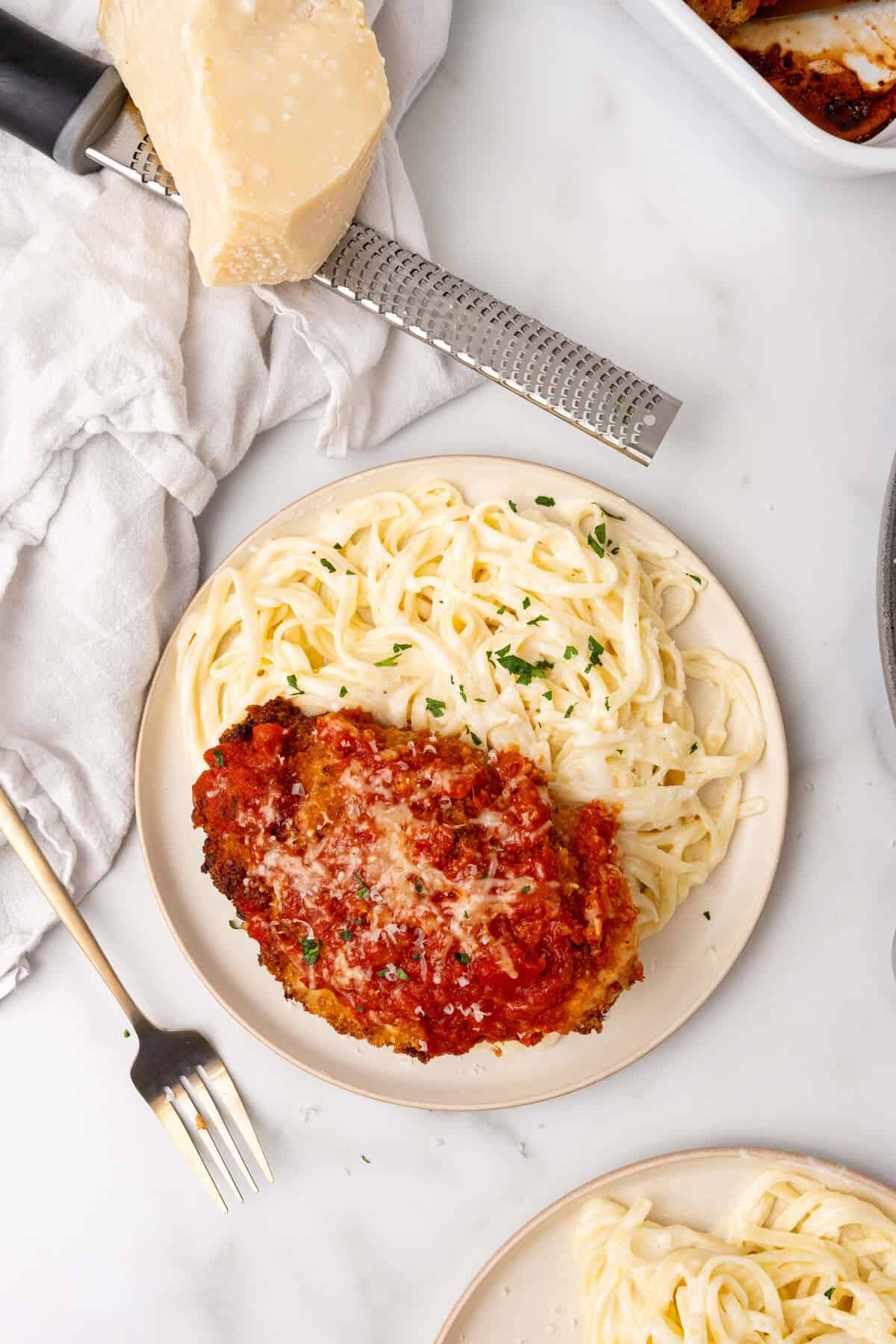 Plate of spaghetti with a Chicken Parmesan Alfredo cutlet and tomato sauce garnished with parsley, with a cheese grater and block of cheese in the background.