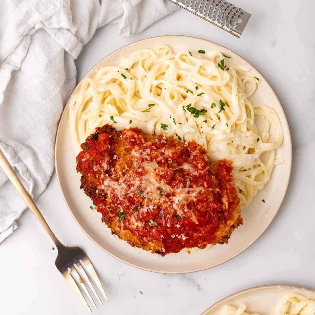 A plate of spaghetti topped with Alfredo sauce next to a breaded Chicken Parmesan cutlet with marinara sauce and grated cheese, garnished with parsley.