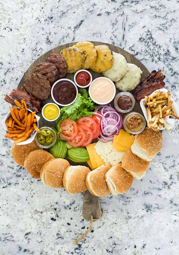 A platter of assorted foods including burgers, steaks, fries, and condiments on a marble surface.