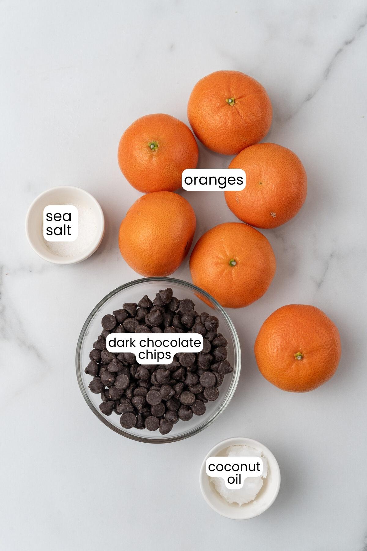 A top-down view of ingredients: chocolate covered oranges, dark chocolate chips, coconut oil, and sea salt on a marble surface, each labeled accordingly.