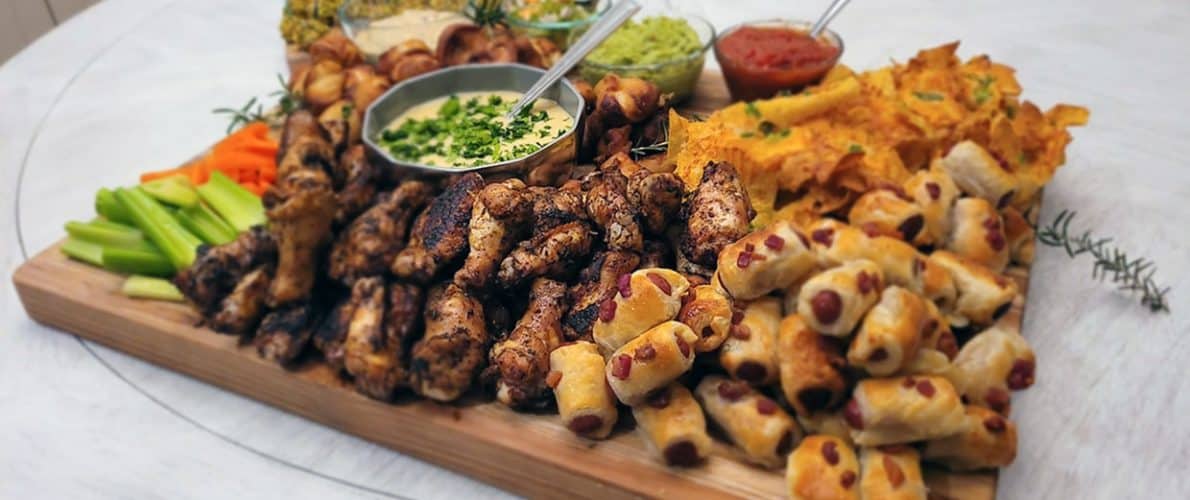 A wooden platter with an assortment of appetizers including grilled chicken skewers, veggie sticks, dips, and pastry-wrapped cocktail sausages.