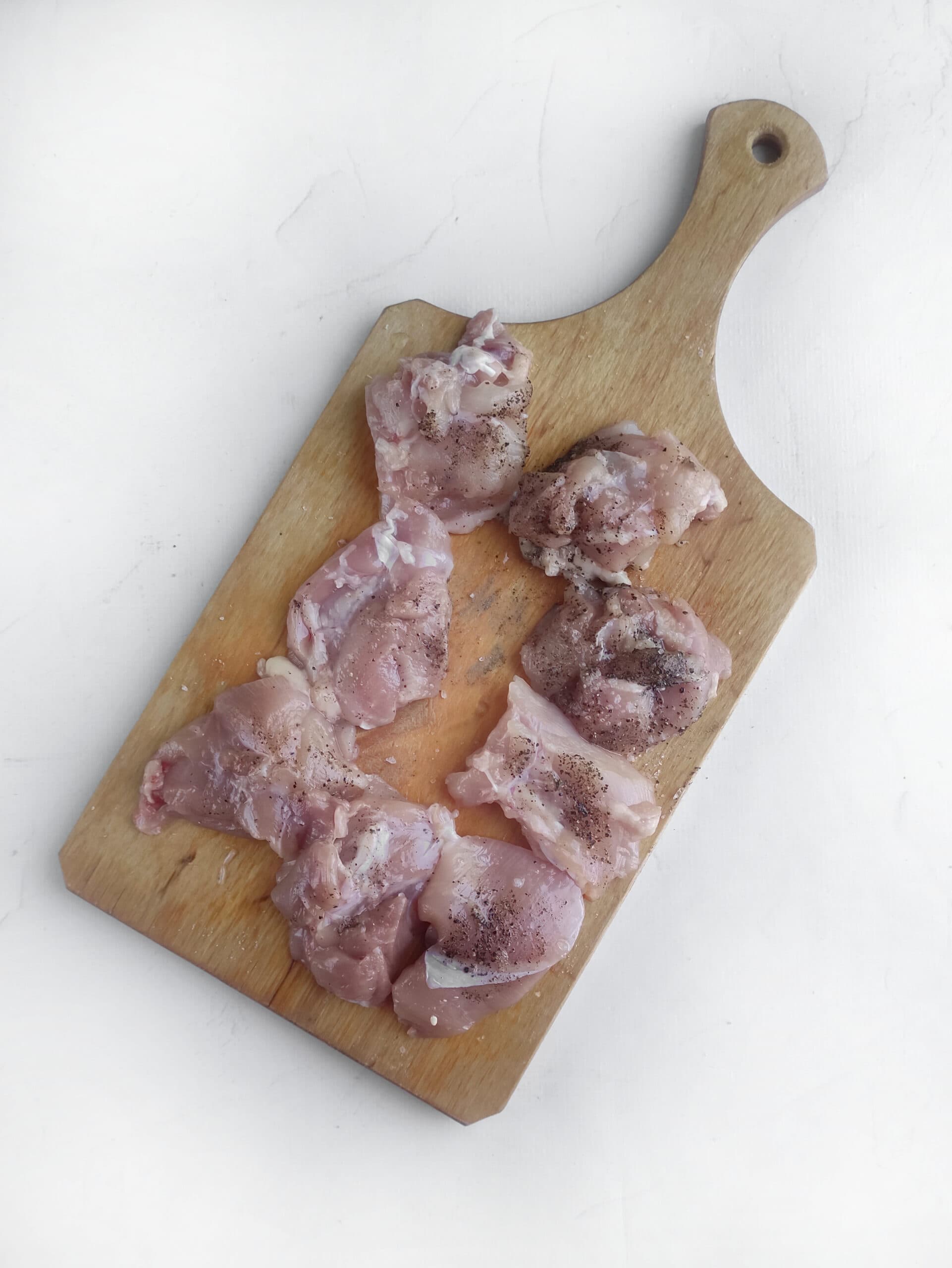 Raw chicken pieces on a wooden cutting board, seasoned with salt and pepper, ready to transform into a delectable Cashew Chicken dish, placed on a white surface.