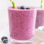A glass of pink berry smoothie, made with almond milk and adorned with a green-striped straw, is topped with blueberries and sits on a white plate.