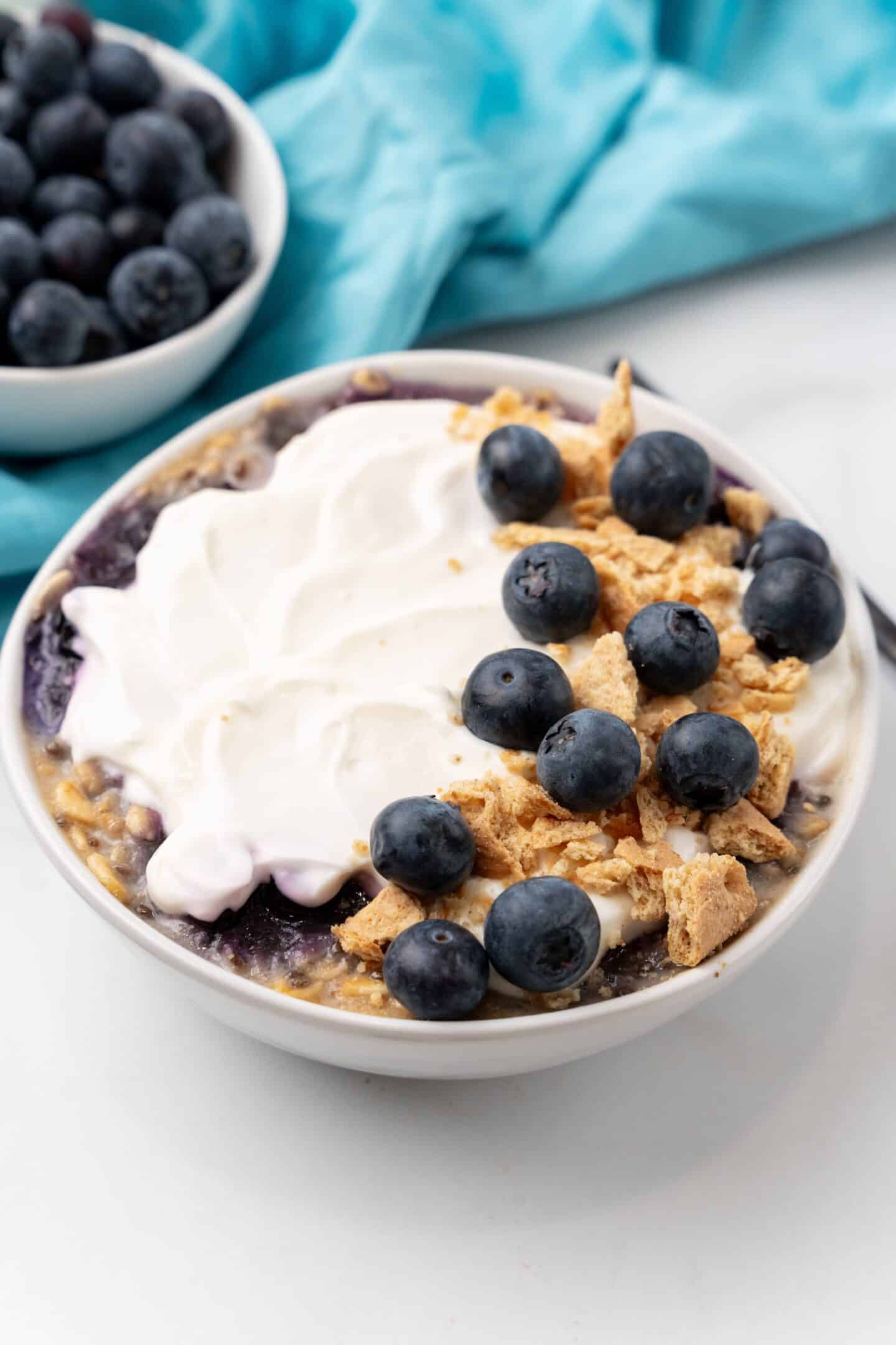 A bowl of creamy yogurt, reminiscent of blueberry cheesecake, is topped with crunchy granola crumbs and fresh blueberries. Nearby, a dish overflows with vibrant berries against a soft blue cloth backdrop.