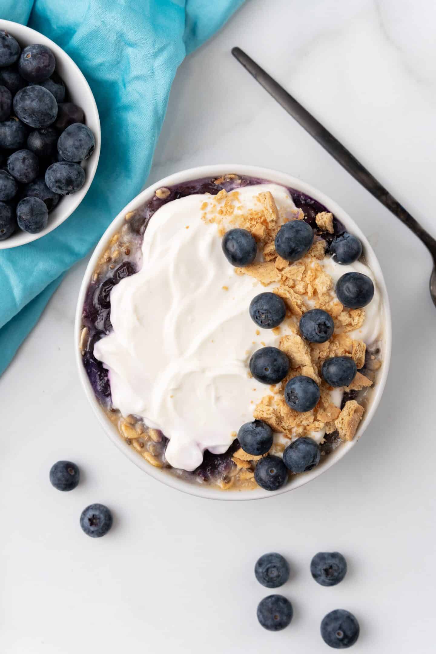 A bowl of yogurt, reminiscent of a blueberry cheesecake, is garnished with granola and fresh blueberries. It's elegantly set on a marble surface beside a spoon and extra blueberries in a small dish, with the edge of a blue cloth peeking into the scene.