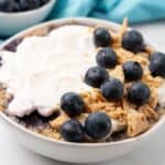 A bowl of yogurt topped with fresh blueberries and crumbled granola sits on a table, evoking the sweet allure of blueberry cheesecake. Nearby, a small bowl of extra blueberries adds the finishing touch to this tempting treat.