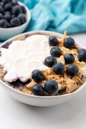 A bowl of yogurt topped with fresh blueberries and crumbled granola sits on a table, evoking the sweet allure of blueberry cheesecake. Nearby, a small bowl of extra blueberries adds the finishing touch to this tempting treat.