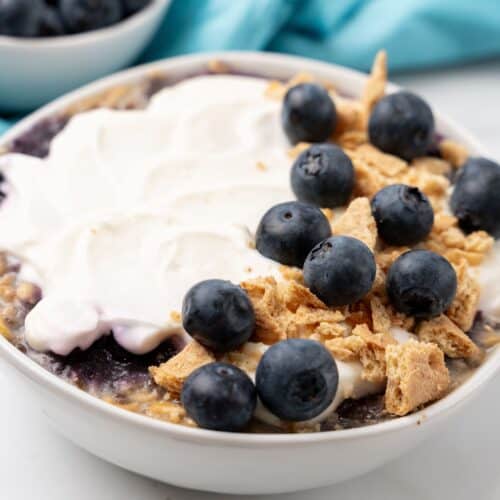 A bowl of yogurt topped with fresh blueberries and crumbled granola sits on a table, evoking the sweet allure of blueberry cheesecake. Nearby, a small bowl of extra blueberries adds the finishing touch to this tempting treat.