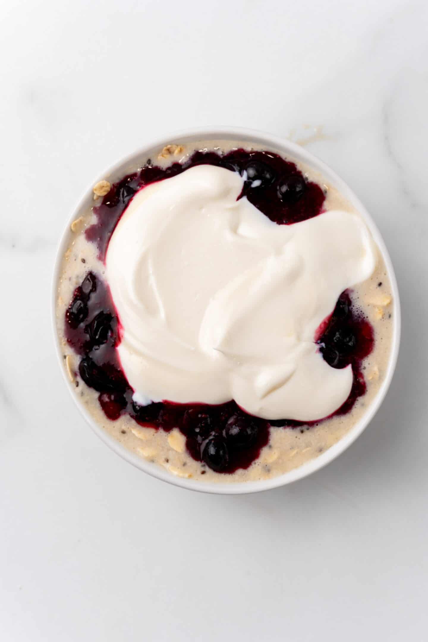 A bowl of overnight oats adorned with dark berries and a dollop of creamy yogurt, reminiscent of a blueberry cheesecake, set against a pristine white background.