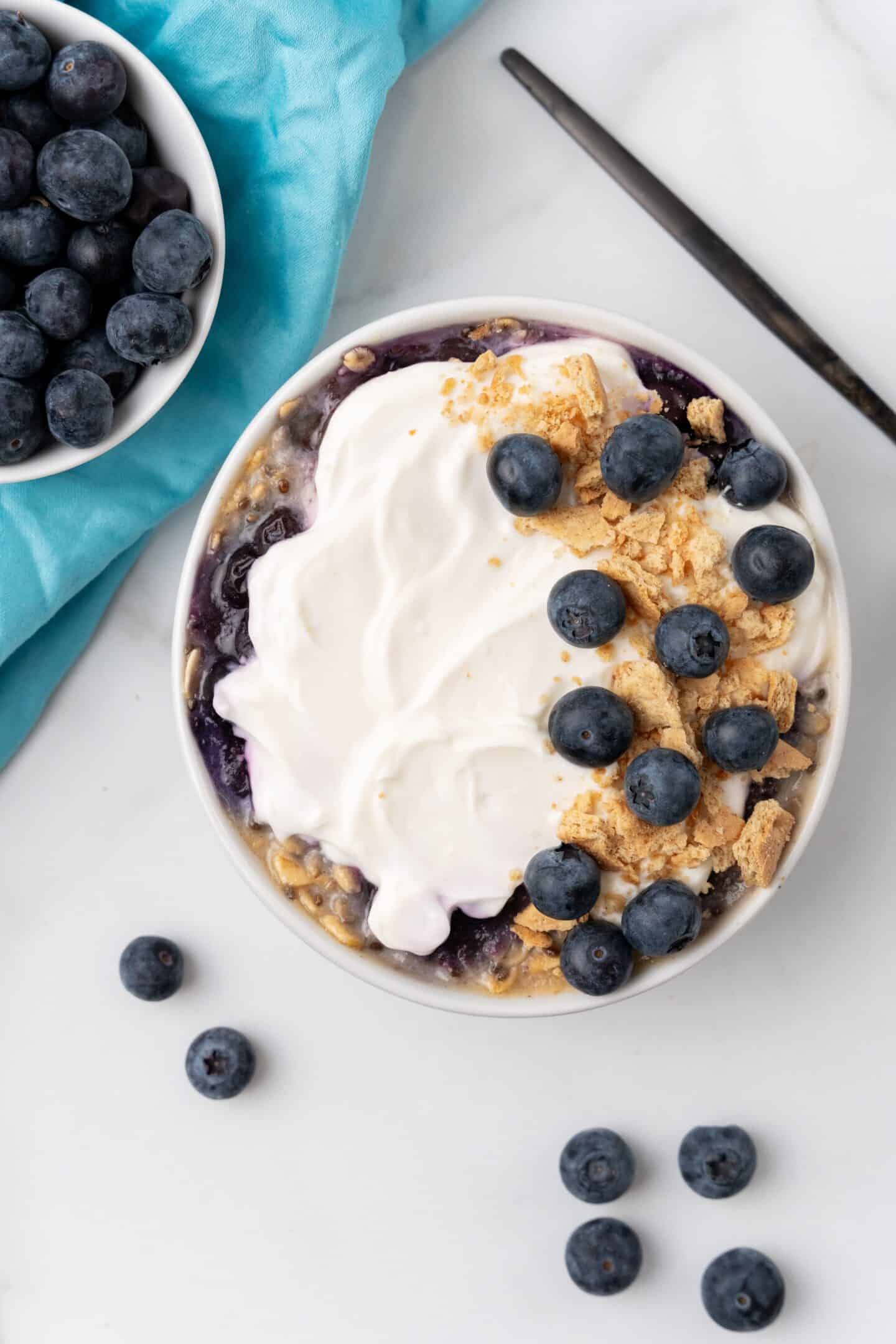 A bowl of creamy yogurt topped with blueberries, granola, and a drizzle of honey sits invitingly next to a blue napkin and spoon. Additional blueberries are scattered on the marble surface, enhancing this enticing breakfast recipe with a hint of blueberry cheesecake allure.