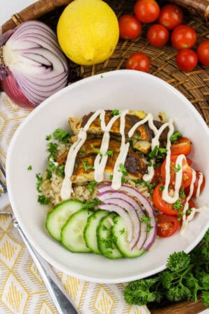 Savor the vibrant Chicken Shawarma Bowl featuring grilled chicken, brown rice, cucumber, red onion, and cherry tomatoes topped with a drizzle sauce. Complemented by fresh lemon, cherry tomatoes, red onion, and parsley on a woven basket for an enticing touch.