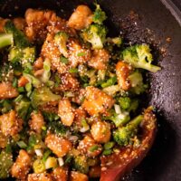 This Honey Garlic Chicken recipe features a delightful stir-fry dish in a pan, combining tender chicken, fresh broccoli, sesame seeds, and green onions. Everything is perfectly mixed with a wooden spoon to ensure every bite is flavorful.