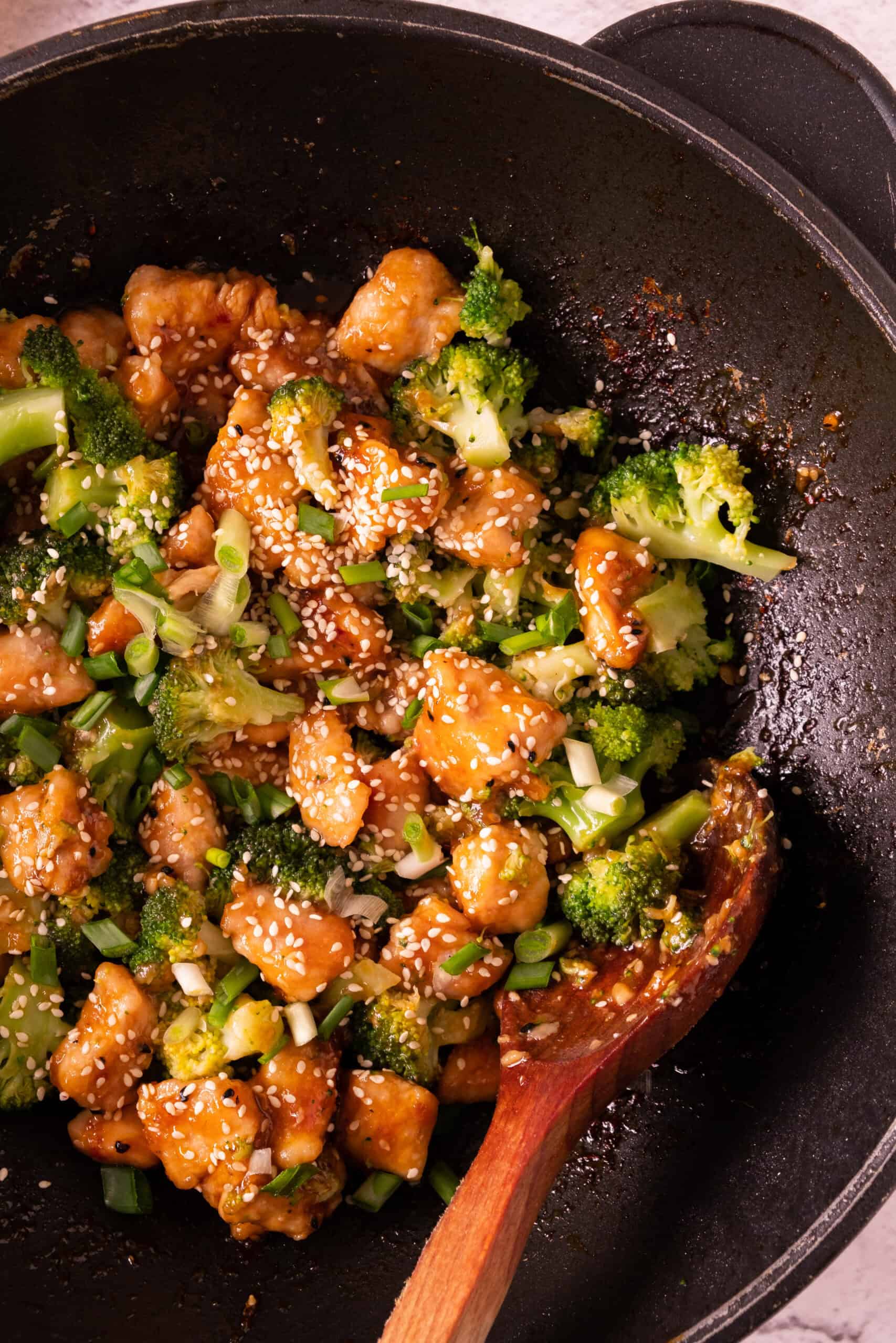 This Honey Garlic Chicken recipe features a delightful stir-fry dish in a pan, combining tender chicken, fresh broccoli, sesame seeds, and green onions. Everything is perfectly mixed with a wooden spoon to ensure every bite is flavorful.