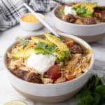 A bowl of taco soup, is topped with avocado slices, sour cream, cheese, and cilantro. A spoon awaits your first bite. In the background, lime wedges and fresh cilantro sit beside a bowl of cheese.