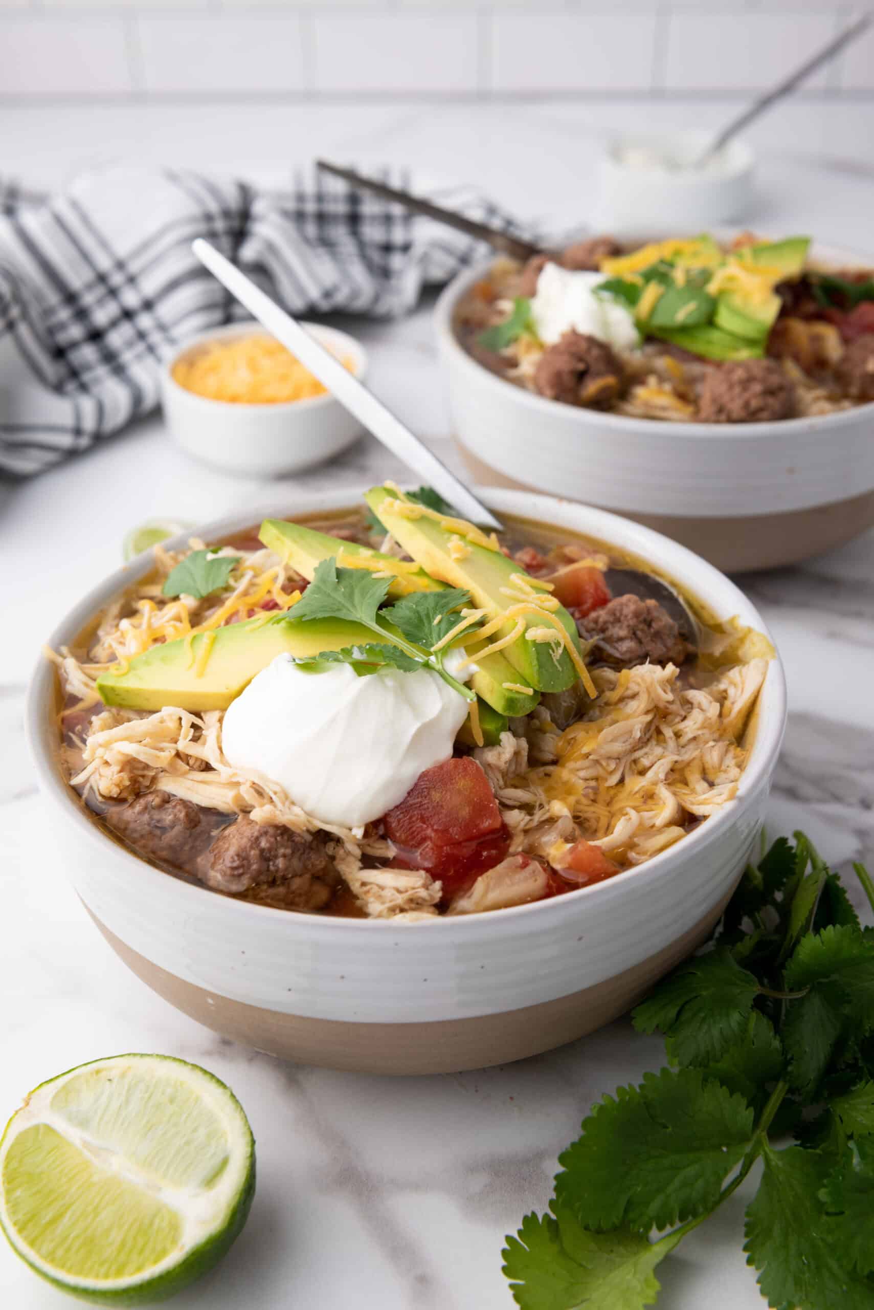 A bowl of taco soup, is topped with avocado slices, sour cream, cheese, and cilantro. A spoon awaits your first bite. In the background, lime wedges and fresh cilantro sit beside a bowl of cheese.