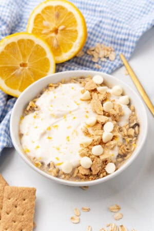 A bowl of lemon cheesecake overnight oats crowned with whipped cream, white chocolate chips, and crumbled cookies, all nestled among fresh lemon slices and a checked cloth.