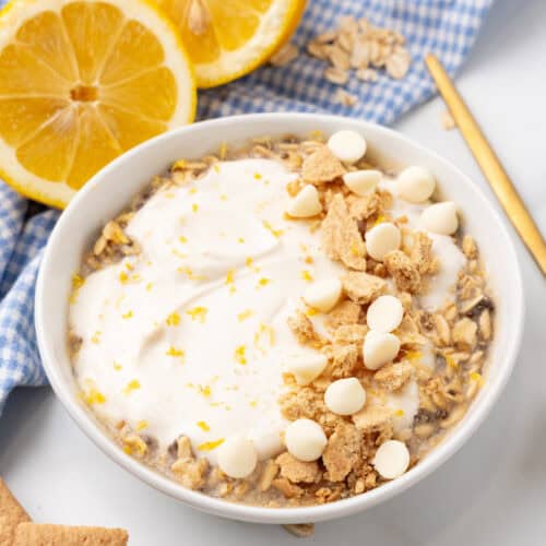 A bowl of lemon cheesecake overnight oats crowned with whipped cream, white chocolate chips, and crumbled cookies, all nestled among fresh lemon slices and a checked cloth.