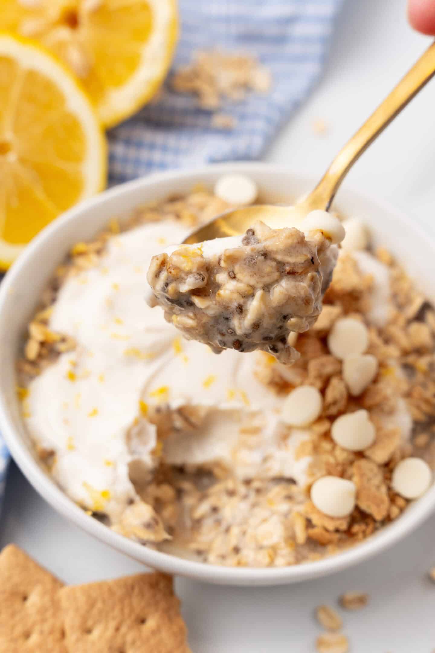 A spoonful of overnight oats with chia seeds, reminiscent of lemon cheesecake, is topped with white chocolate chips and yogurt. It hovers above a bowl, while lemons and a blue cloth add a fresh backdrop.