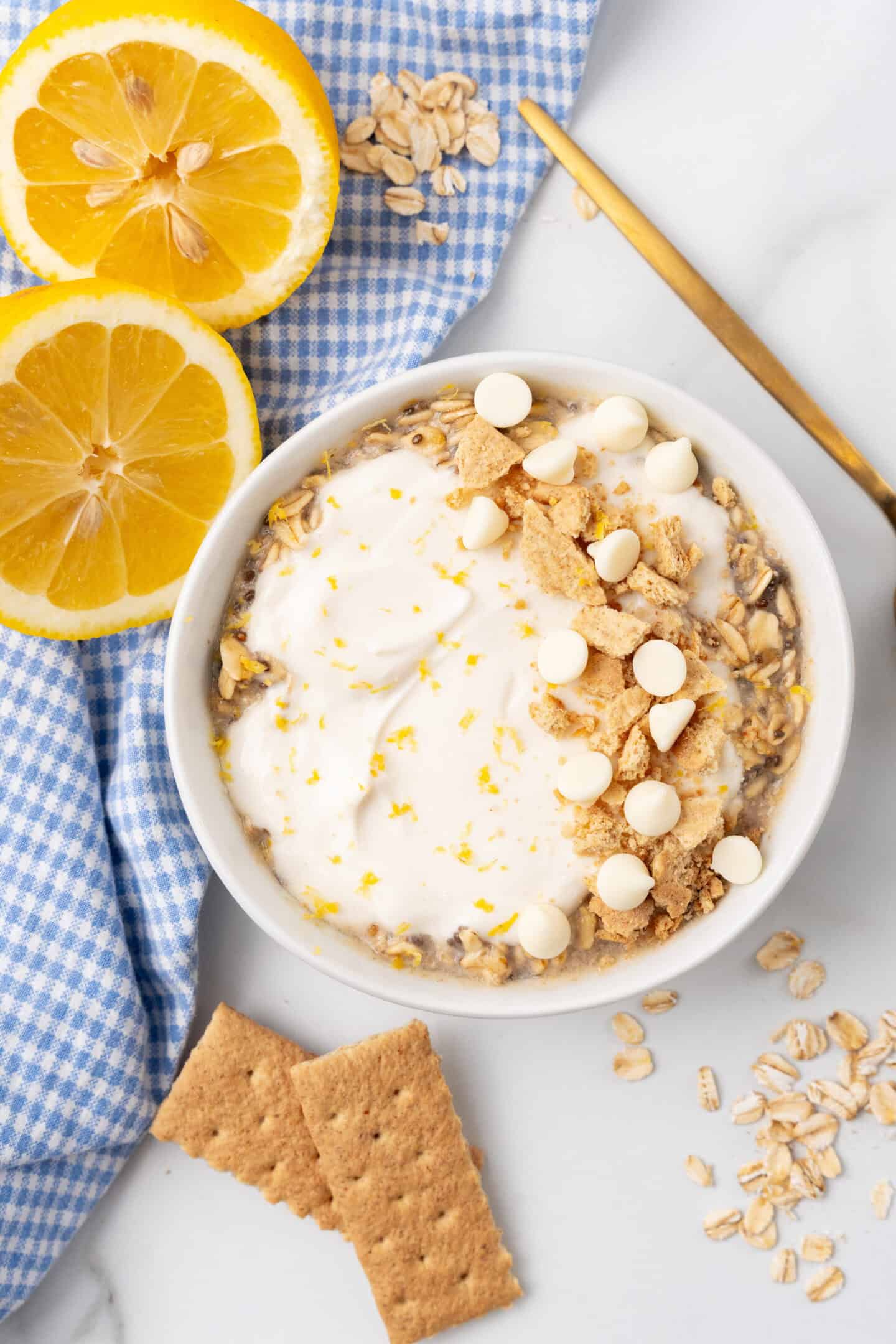 A bowl of overnight oats topped with yogurt, crumbled cookies, and white chocolate chips exudes a delightful lemon cheesecake vibe. Two lemon slices, a blue checkered cloth, and a spoon complete the scene.