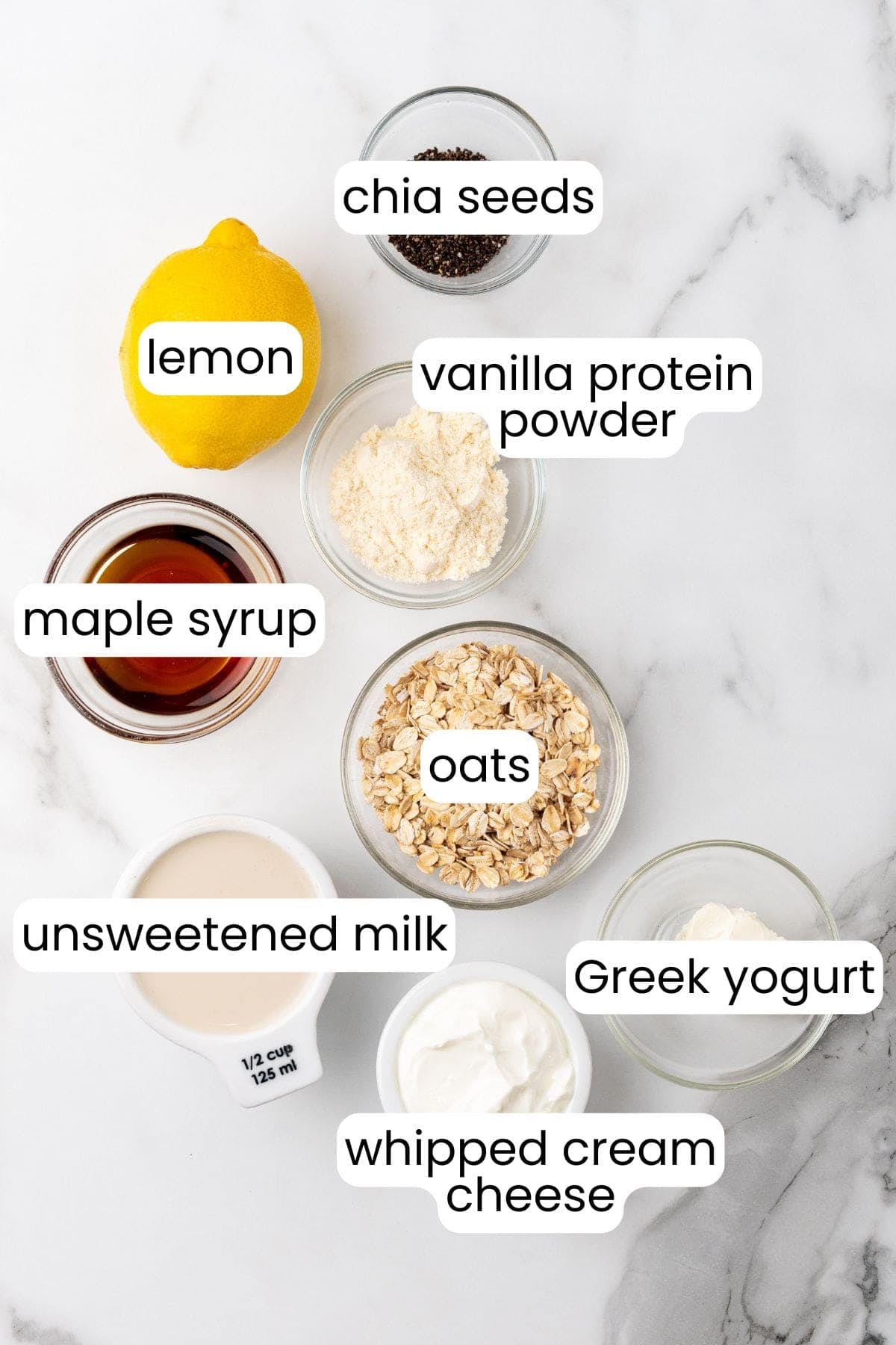 Overhead view of ingredients for Lemon Cheesecake Overnight Oats: lemon, chia seeds, vanilla protein powder, maple syrup, oats, unsweetened milk, Greek yogurt, and whipped cream cheese, each labeled in bowls.