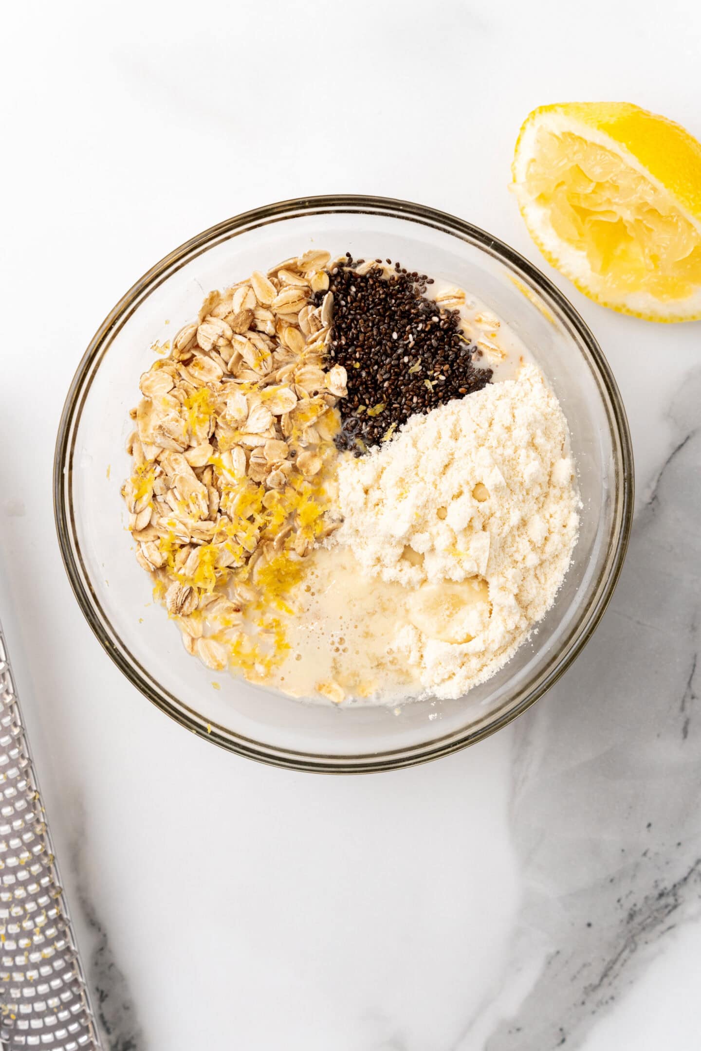 A bowl of overnight oats with chia seeds, almond flour, and lemon zest sits on a marble surface, its flavors reminiscent of a lemon cheesecake. A partially squeezed lemon wedge rests beside it, poised to add just a touch more zing.
