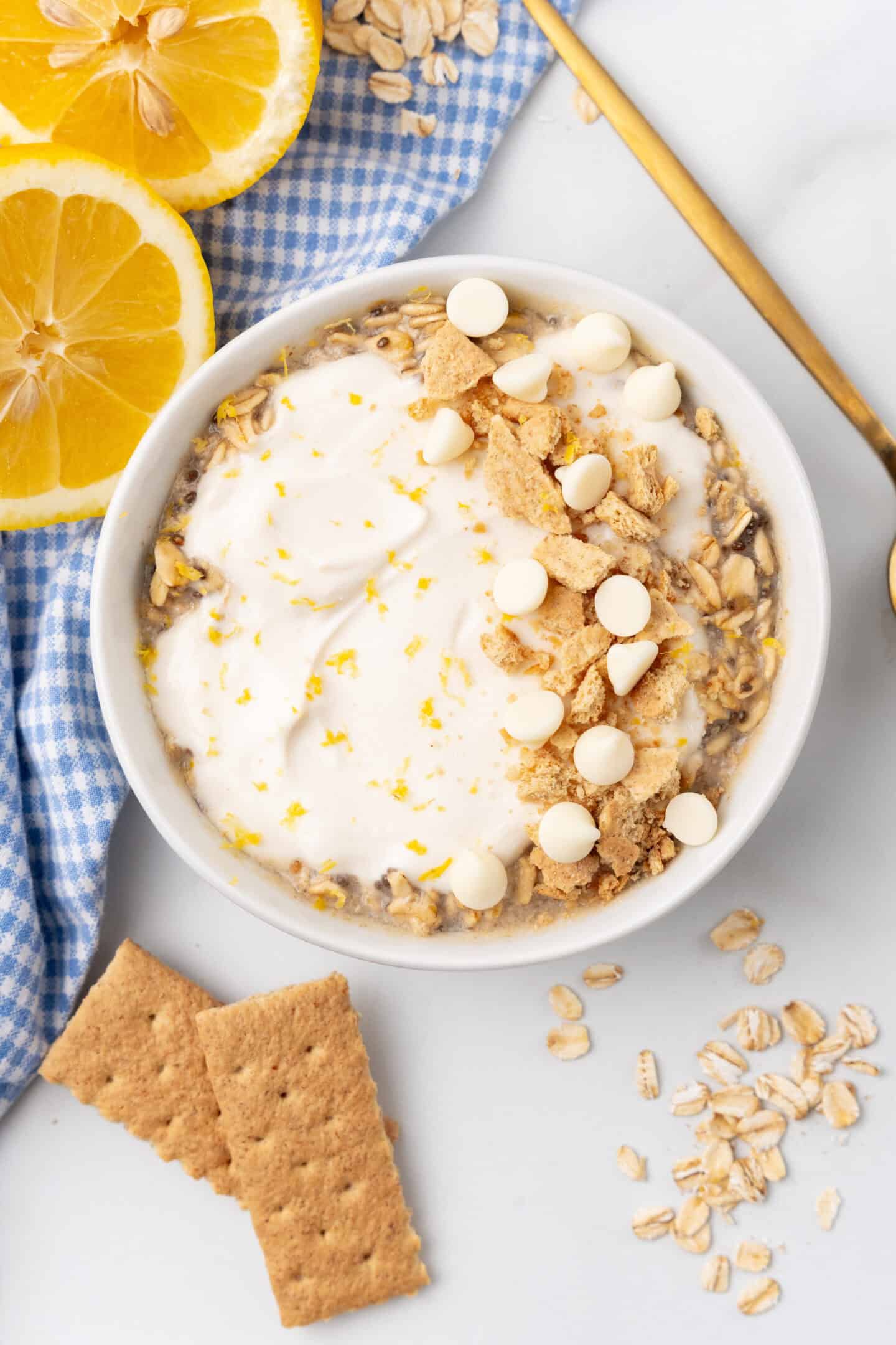 A bowl of yogurt reminiscent of lemon cheesecake, topped with white chocolate chips, crushed graham crackers, and lemon zest. Surrounded by lemon slices, oats for a nod to overnight oats, graham crackers, and a blue checkered cloth.