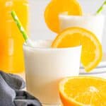 A frothy Orange Julius protein shake in a glass with a green straw, garnished with an orange slice, sits next to a halved orange. A second glass and a bottle of orange juice are visible in the background.
