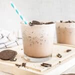 A glass filled with a cookies and cream protein shake topped with crushed Oreos, accompanied by a second glass in the background. A blue and white striped straw is in the glass.