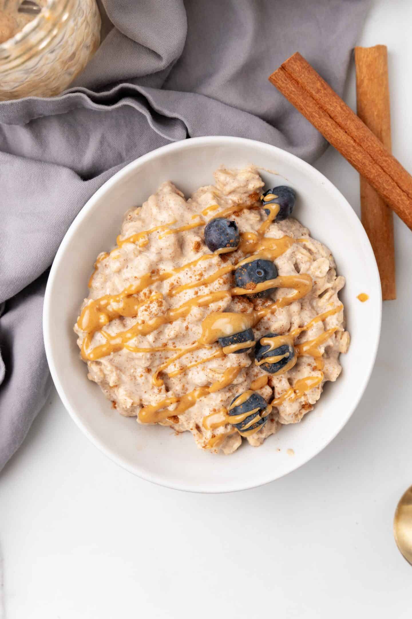 A bowl of overnight oats topped with blueberries and drizzled with peanut butter, placed on a gray cloth beside two cinnamon sticks.