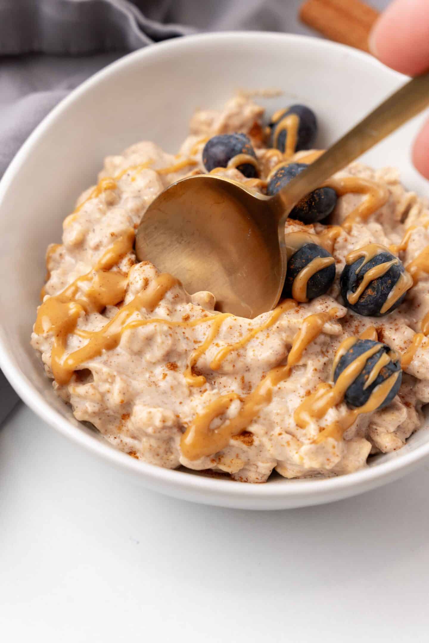 A bowl of creamy overnight oats with a delightful peanut butter drizzle, topped with juicy blueberries and a sprinkle of chia seeds, being gently stirred with a spoon.